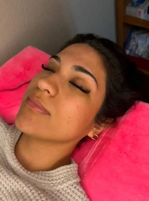 Woman resting on pink pillow with eyes closed, showing eyelash extensions and groomed eyebrows